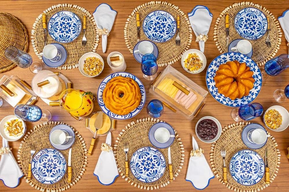 Mesa de praia posta com sousplat de palha e búzios, louça azul e branca e talheres de bambu